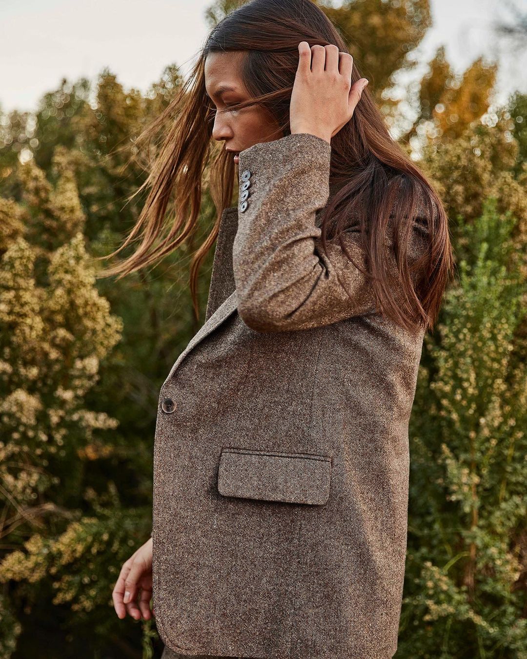 Adult woman with long brown hair poses in a brown tweed blazer outdoors against a blurred background of green trees and vegetation.