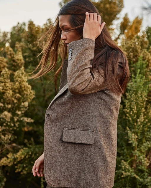 Adult woman with long brown hair poses in a brown tweed blazer outdoors against a blurred background of green trees and vegetation.