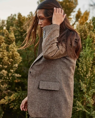 Adult woman with long brown hair poses in a brown tweed blazer outdoors against a blurred background of green trees and vegetation.