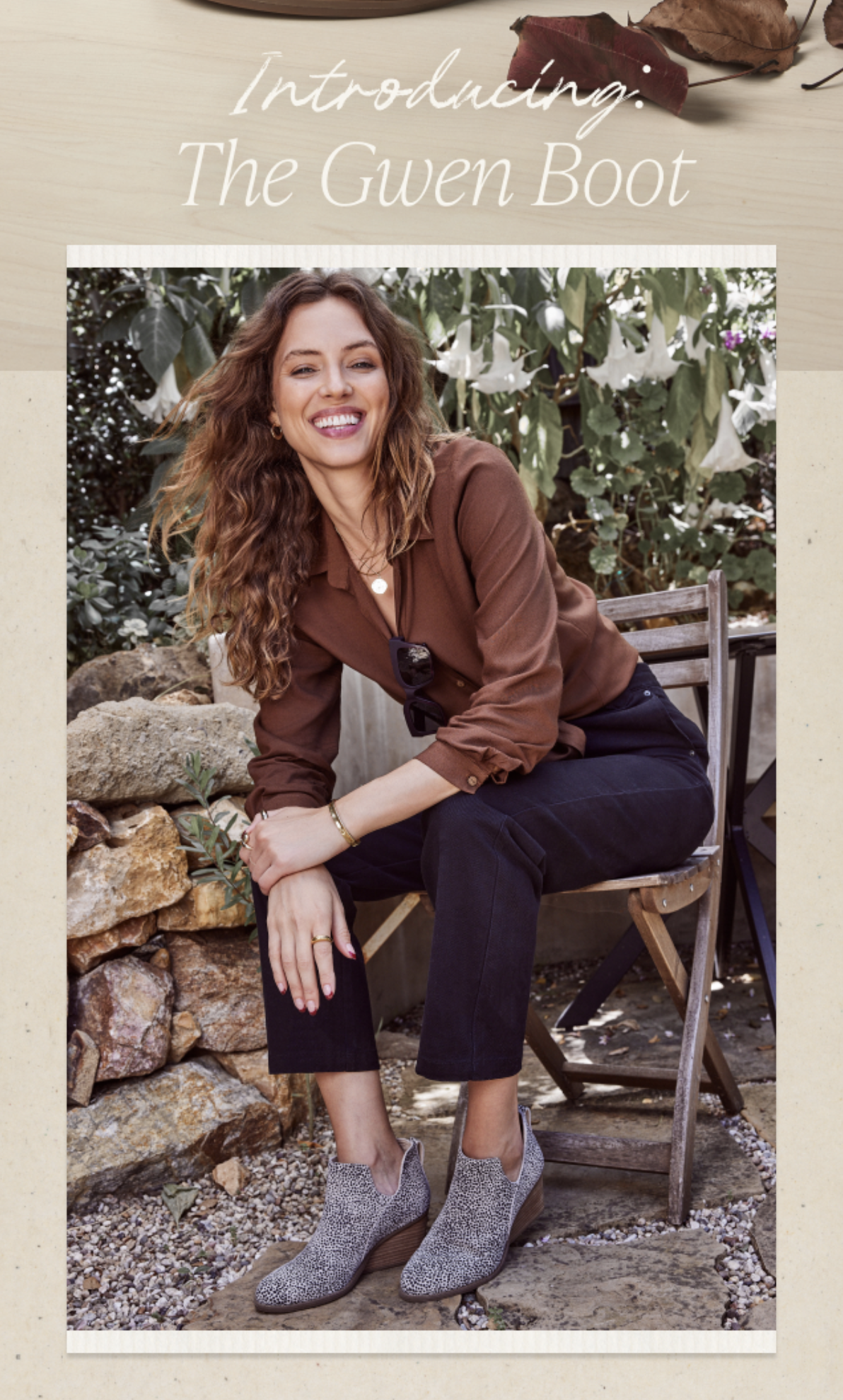 Smiling woman in a brown blouse and black pants sits on a wooden chair wearing grey speckled ankle boots in an outdoor setting.