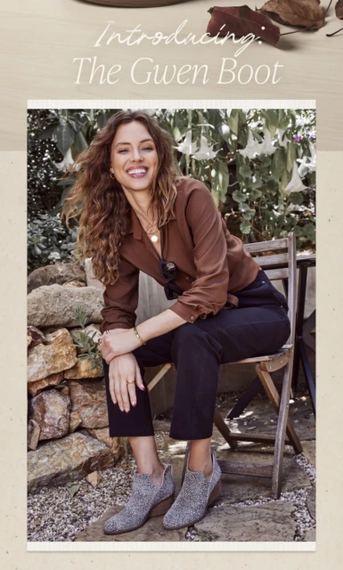 Smiling woman in a brown blouse and black pants sits on a wooden chair wearing grey speckled ankle boots in an outdoor setting.