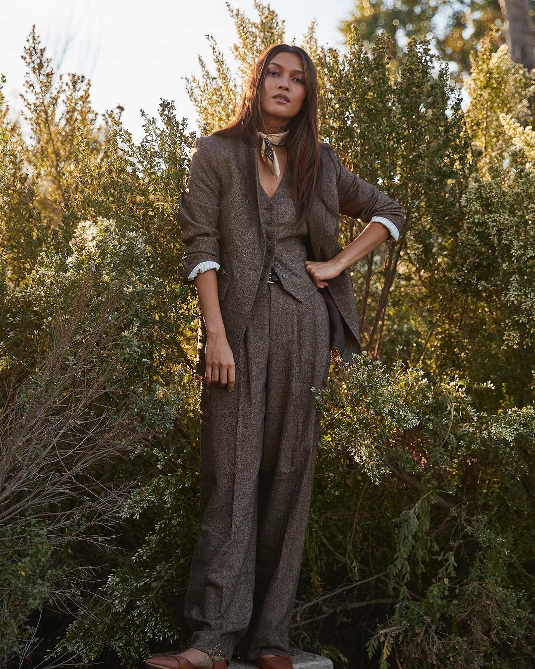 A woman wearing a brown tweed pantsuit with a tan scarf stands outdoors surrounded by green shrubs in natural lighting.