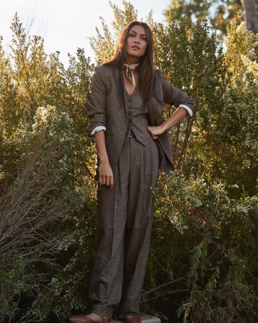 A woman wearing a brown tweed pantsuit with a tan scarf stands outdoors surrounded by green shrubs in natural lighting.