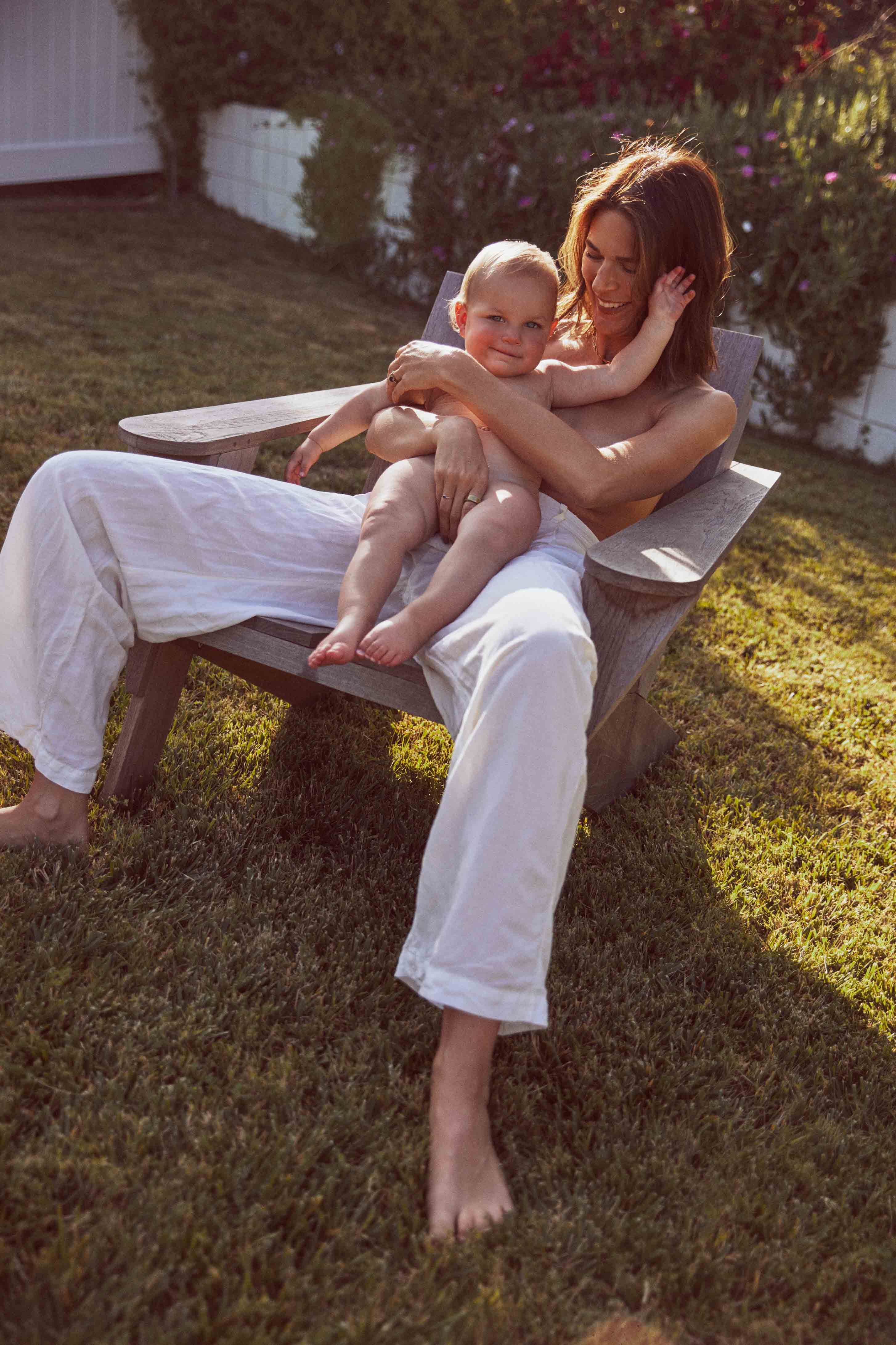 A woman sits outside in a wood chair, barefoot, holding a young child in her lap on a sunny day surrounded by a lawn and greenery.
