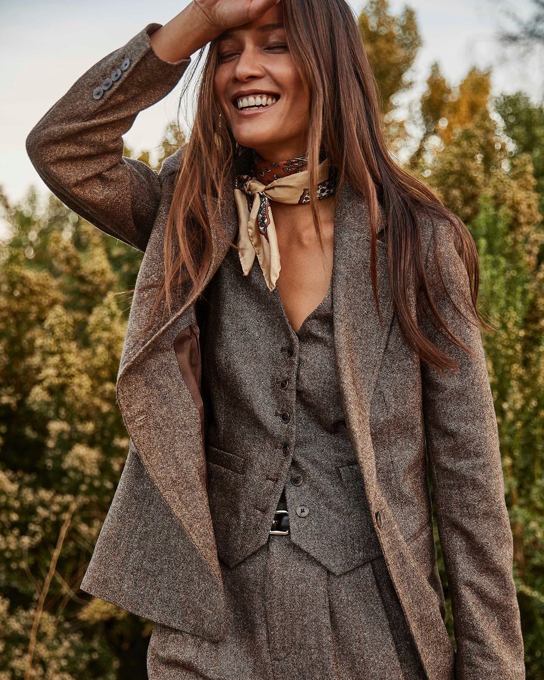 Smiling woman in tweed suit stands outdoors with trees in soft-focus background. She wears a patterned scarf. Autumn fashion.