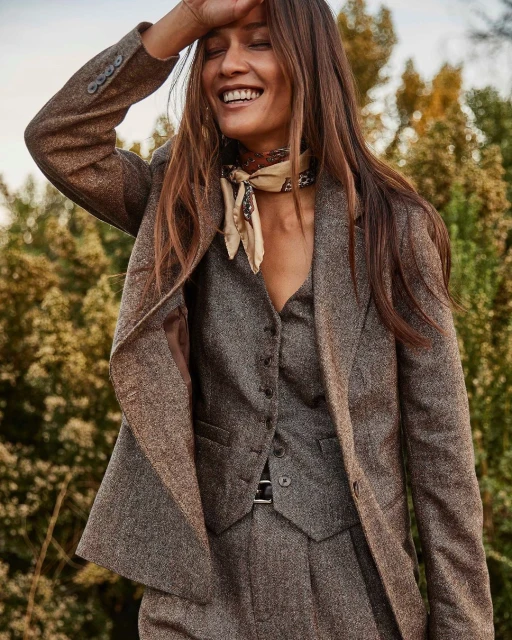 Smiling woman in tweed suit stands outdoors with trees in soft-focus background. She wears a patterned scarf. Autumn fashion.