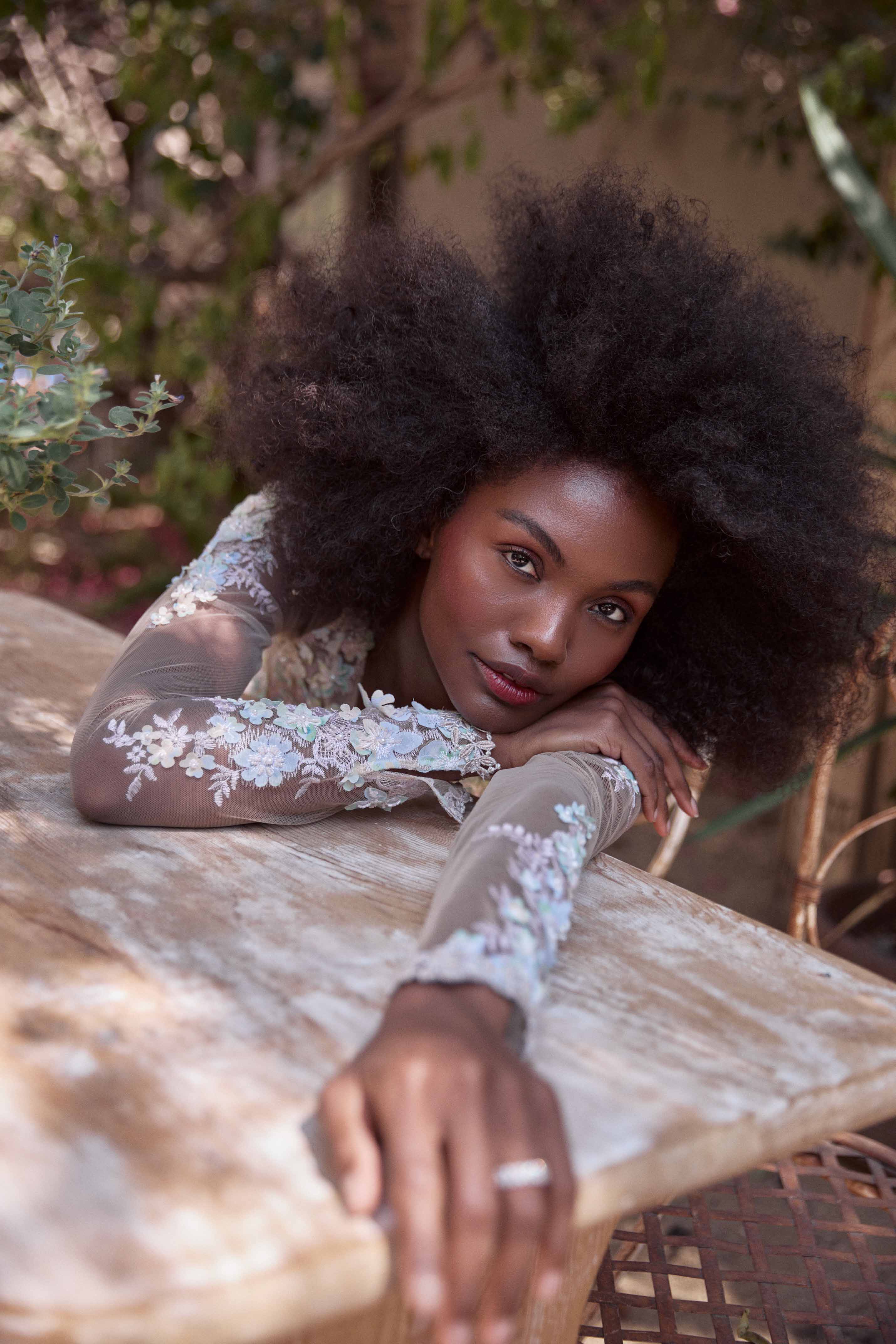 Black woman with voluminous afro hair resting arms on a rustic wooden outdoor table, wearing a sheer floral embroidered top.