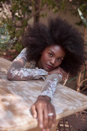 Black woman with voluminous afro hair resting arms on a rustic wooden outdoor table, wearing a sheer floral embroidered top.