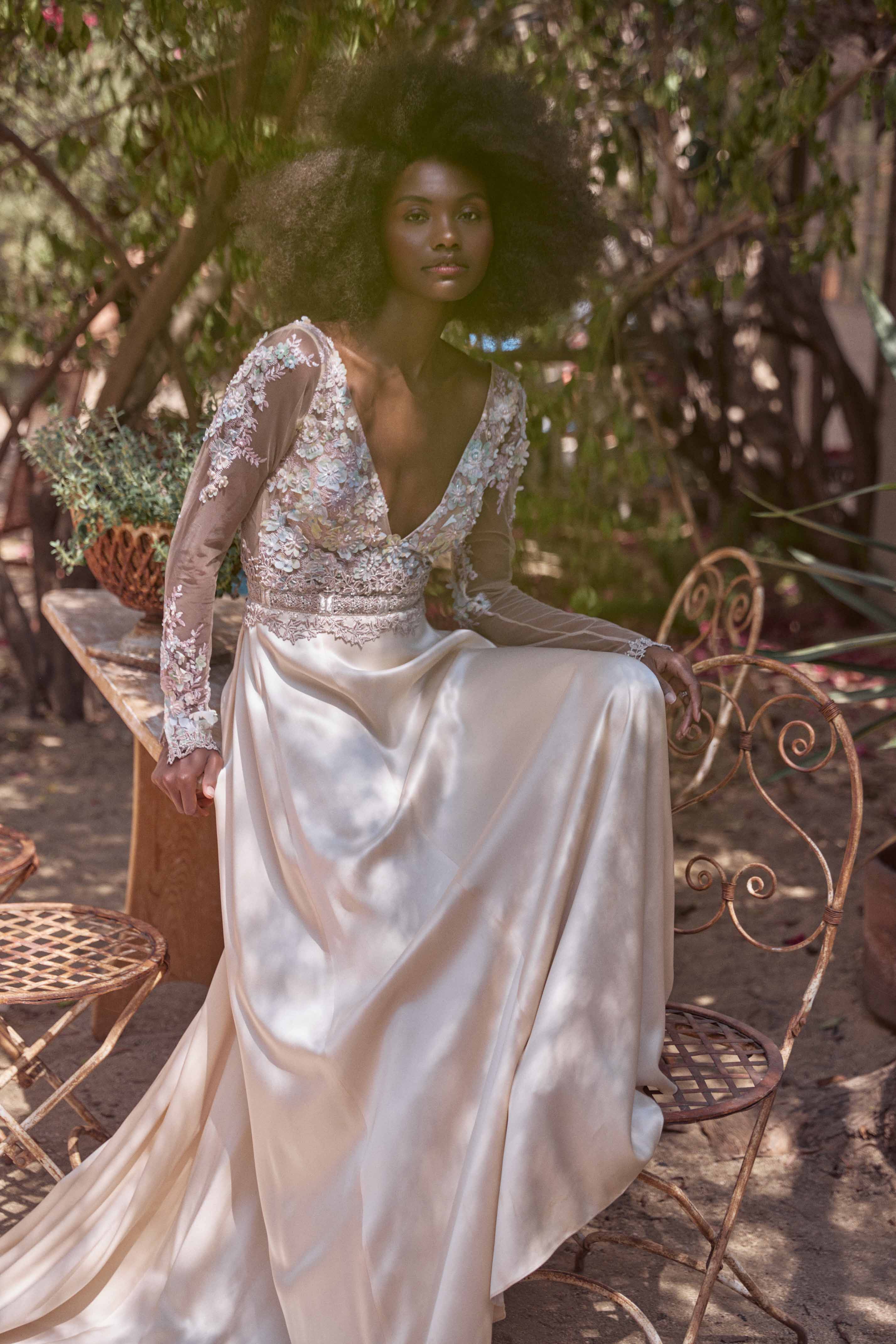 Black woman with voluminous afro hair wearing an off-white silk and lace gown seated on an ornate metal chair outdoors.
