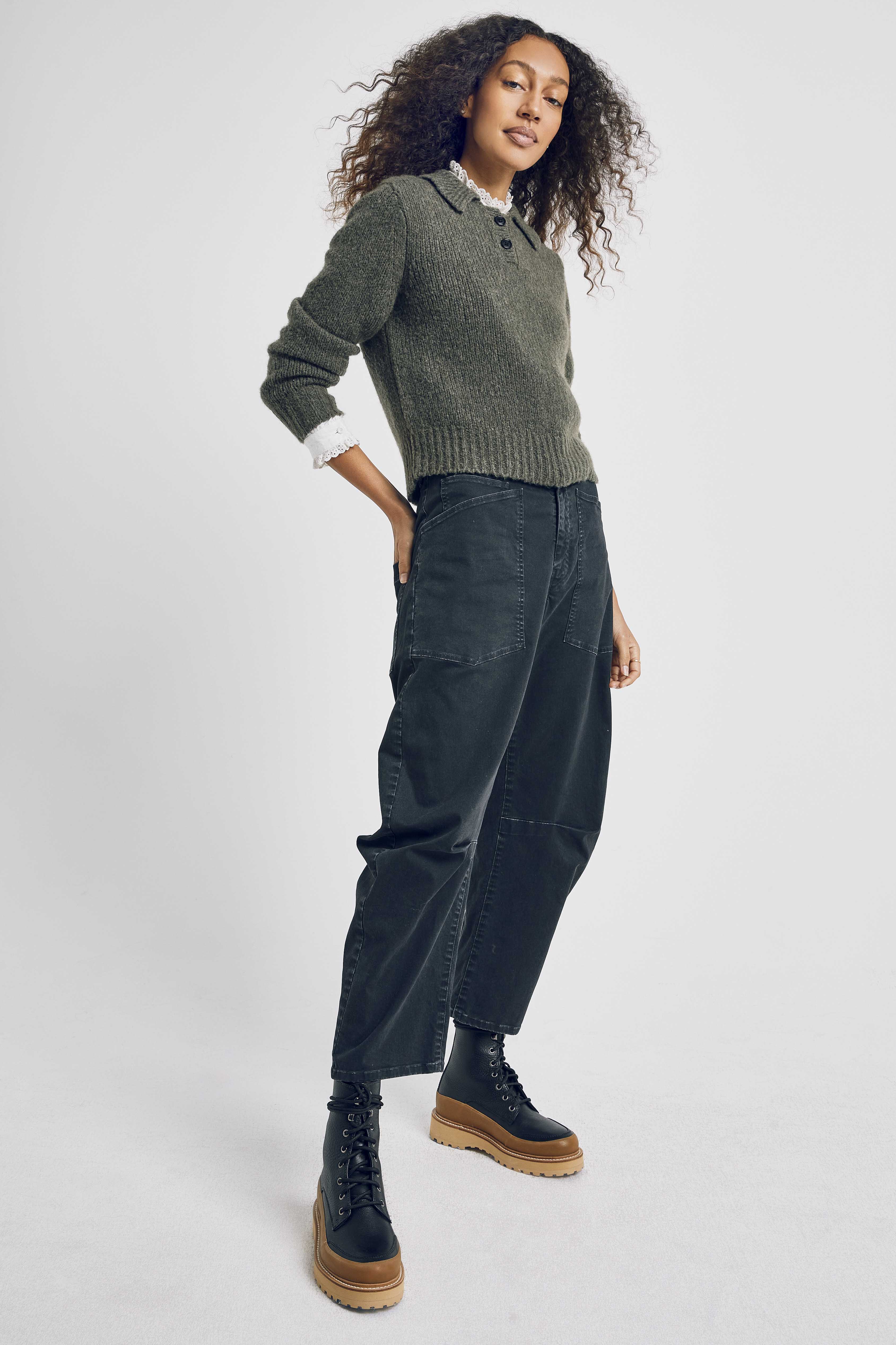 Full-length shot of a woman with curly hair wearing an olive knit sweater, jeans, and platform boots on a white background.
