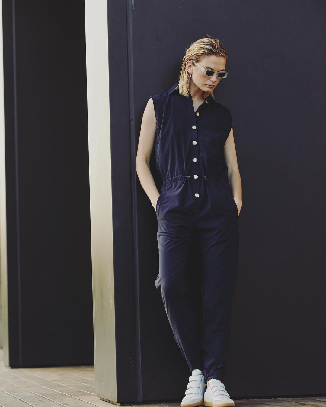 Woman in dark jumpsuit and sunglasses leaning against a dark wall photographed by Annie Edmonds