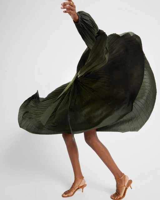 Woman in flowing green dress and strappy heels twirling in a bright studio, showcasing fashion and movement in a dynamic pose.