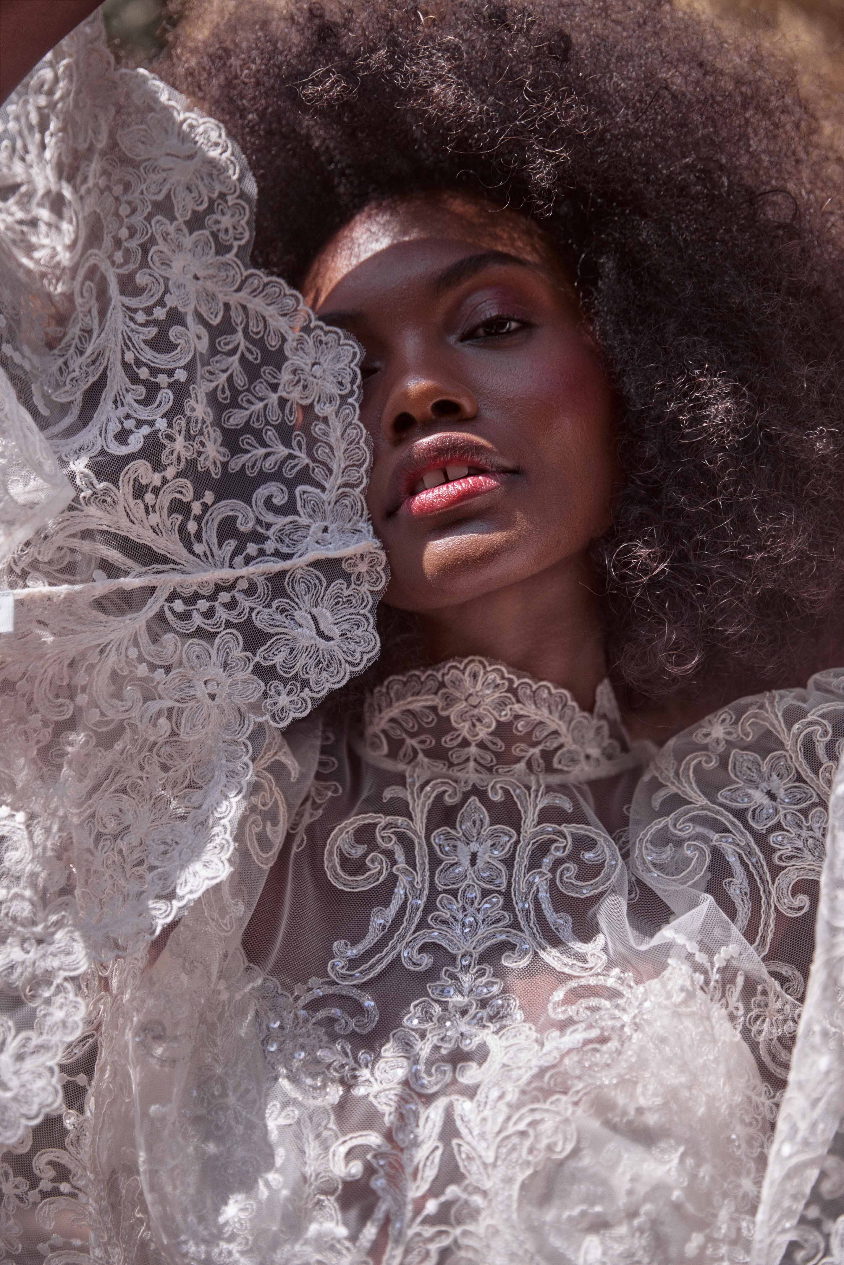 Close-up portrait of a Black woman with large afro, wearing a white lace gown and posing outdoors in natural lighting.