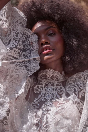 Close-up portrait of a Black woman with large afro, wearing a white lace gown and posing outdoors in natural lighting.
