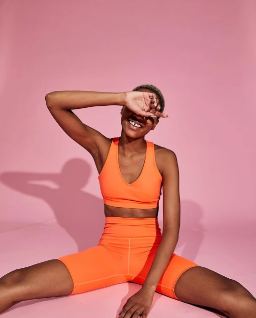 Smiling woman in orange workout clothes covers her face sitting against a pink background in a studio, creating a playful and energetic visual.
