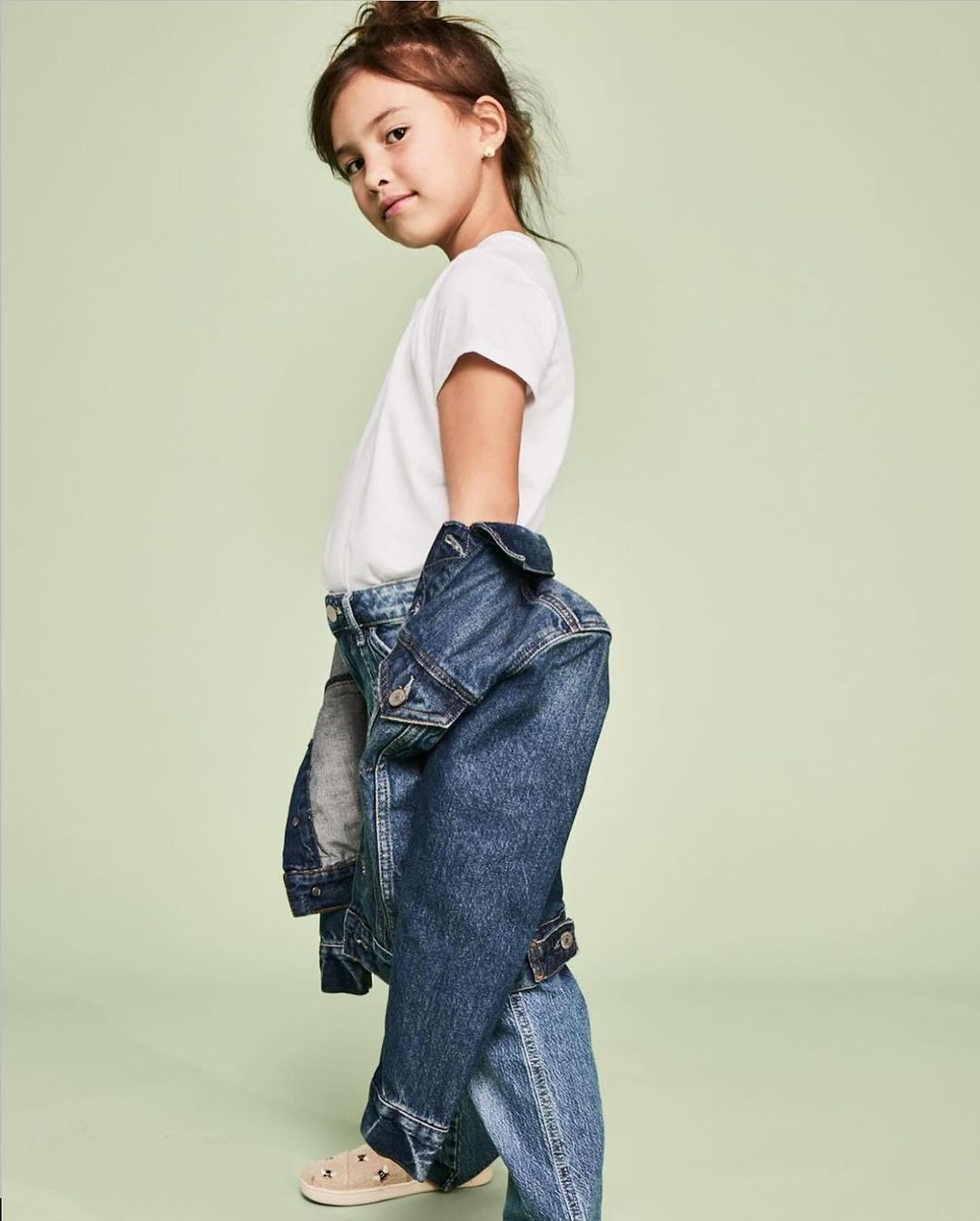 Young girl wearing denim jacket and jeans against a light green backdrop in a studio fashion shot, casual style, looking at the camera.