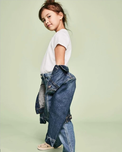 Young girl wearing denim jacket and jeans against a light green backdrop in a studio fashion shot, casual style, looking at the camera.