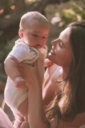 Portrait of a woman holding an infant in a light-colored romper outdoors in natural sunlight, captured by Annie Edmonds.