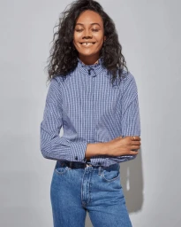 Smiling woman with dark curly hair wearing a blue gingham blouse and blue jeans against a light grey background.