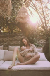 Woman seated outdoors holding a baby on an outdoor daybed beneath woven pendant lights and lush foliage.