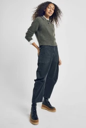 Full-body portrait of a woman with curly hair wearing an olive green sweater, dark utility pants, and platform boots against a white background.