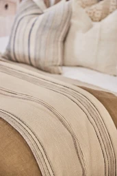 Close-up interior photograph of a neatly made bed with neutral-toned linen bedding, striped throw pillows, and a textured blanket.