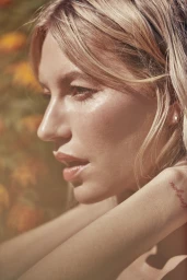 Close-up portrait of a blonde woman with glowing skin outdoors amidst warm, out-of-focus floral background.