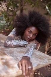 Black woman with voluminous afro hair resting arms on a rustic wooden outdoor table, wearing a sheer floral embroidered top.
