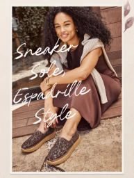 Smiling woman with curly dark hair wearing a brown dress and patterned slip-on shoes while seated on wooden steps.