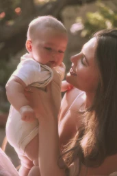 Woman smiling down at an infant baby held in her arms outdoors in soft warm sunlight