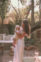 Woman in a long white slip dress holding an infant child in a garden courtyard by Annie Edmonds