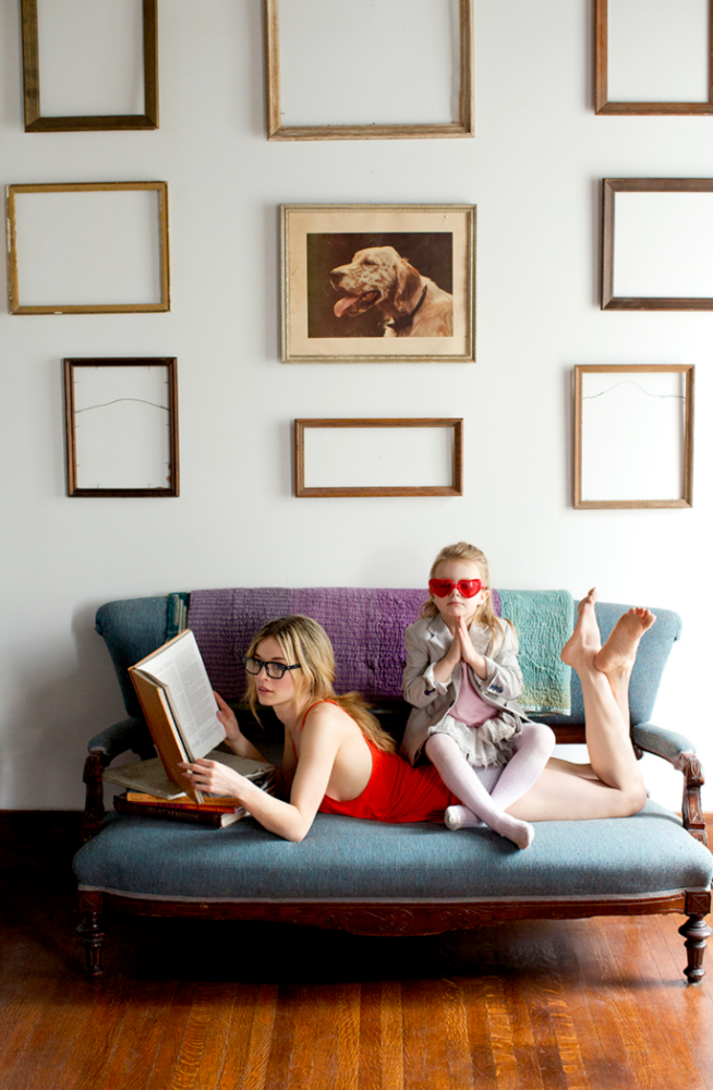 Young adult and child pose together on a teal sofa with wooden floor. Bare wall in the background with decorative picture frames.