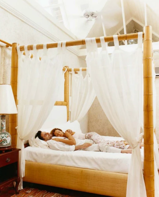 A couple is lying in a four-poster bed, with white sheets and curtains, inside a light and airy bedroom with a lamp on the bedside table.