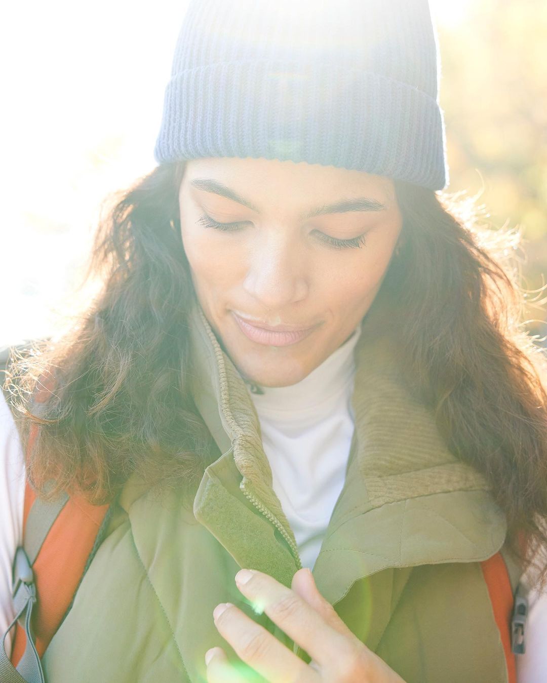 A young adult woman with curly hair wears a blue knit beanie and a green vest while outside in bright sunlight.