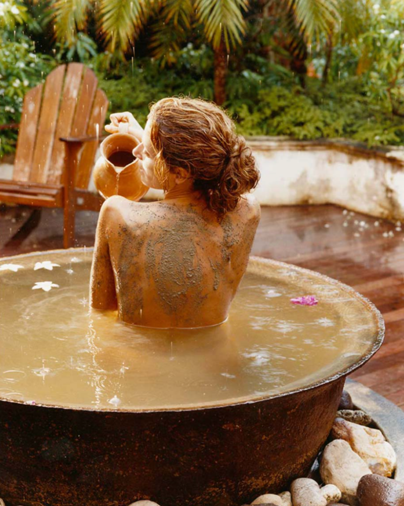 Adult female partially submerged in a round tub outdoors with a body treatment, holding a clay pitcher near a wooden chair and greenery.