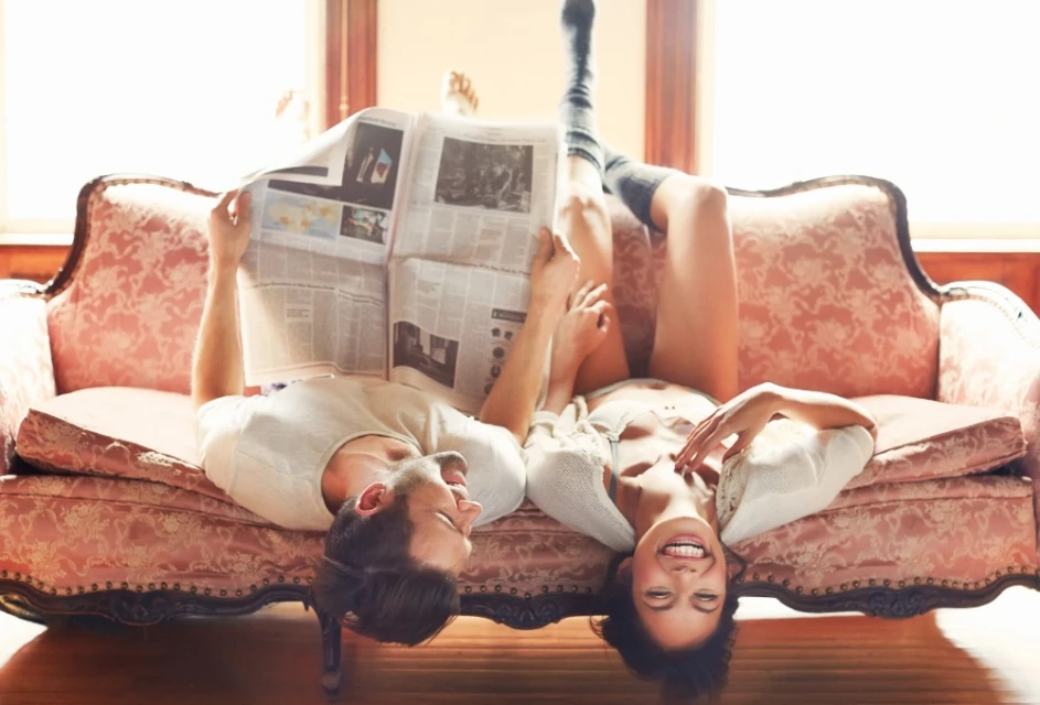 Upside-down indoor shot of a smiling couple lounging on a vintage sofa, the man reading a newspaper and the woman in knee socks.