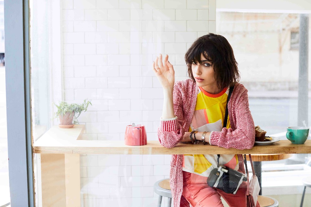 A brunette woman in pink pants sits at a cafe table with a camera and accessories, near a window with city view, captured in a medium shot.