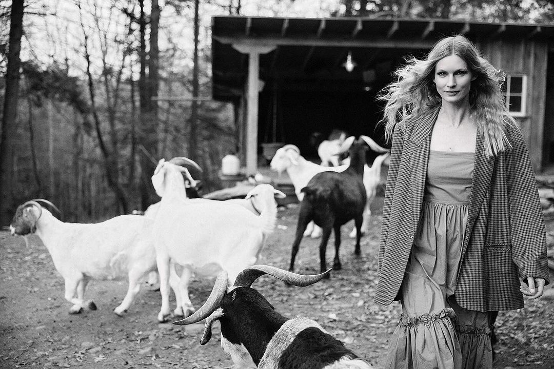 Black and white image of a blonde woman in a blazer standing in a farm yard with a group of goats behind her.