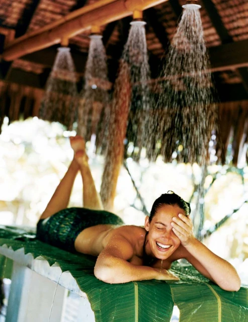Woman relaxing under outdoor showers in tropical spa setting. Lush green banana leaves, thatched roof, and water create serene scene.