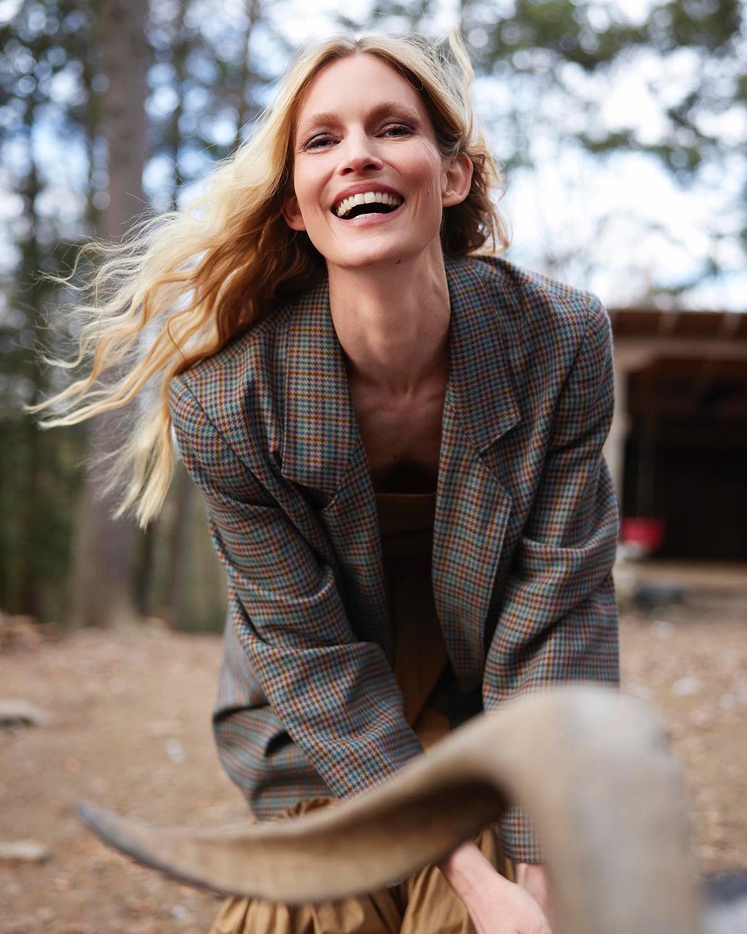 Smiling blonde woman in blazer poses outdoors with a sheep with fleece in the foreground, trees and a structure in the background.