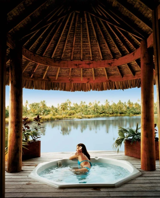 Woman relaxing in a hot tub outdoors, surrounded by a wooden gazebo and lush tropical scenery with a lagoon view.