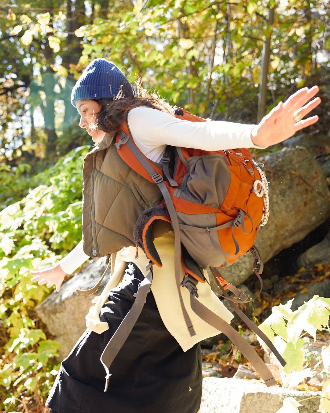 Woman with backpack hiking in a forest, wearing a beanie, vest, and dark skirt, strikes a balancing pose with outstretched arms surrounded by green foliage.