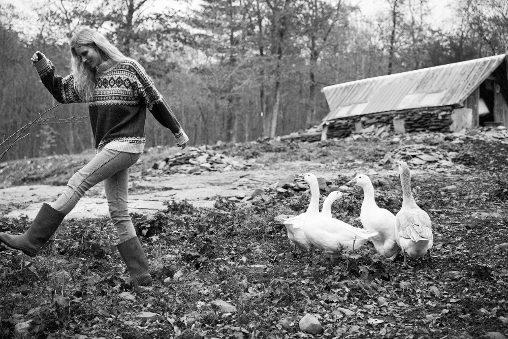 Black and white photo of a woman walking near a flock of white geese in a rural setting with a barn and trees in the background.