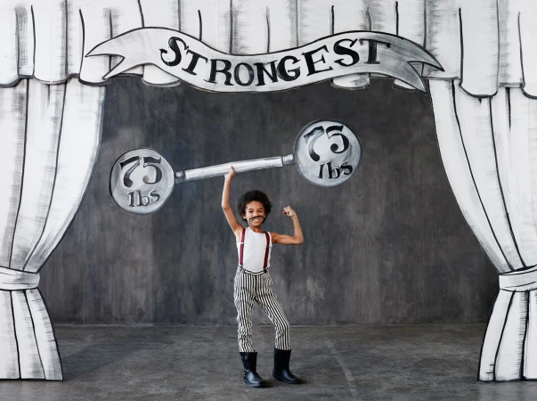Young child in a circus performer costume stands in front of a drawn backdrop with weights and the word 'Strongest'.