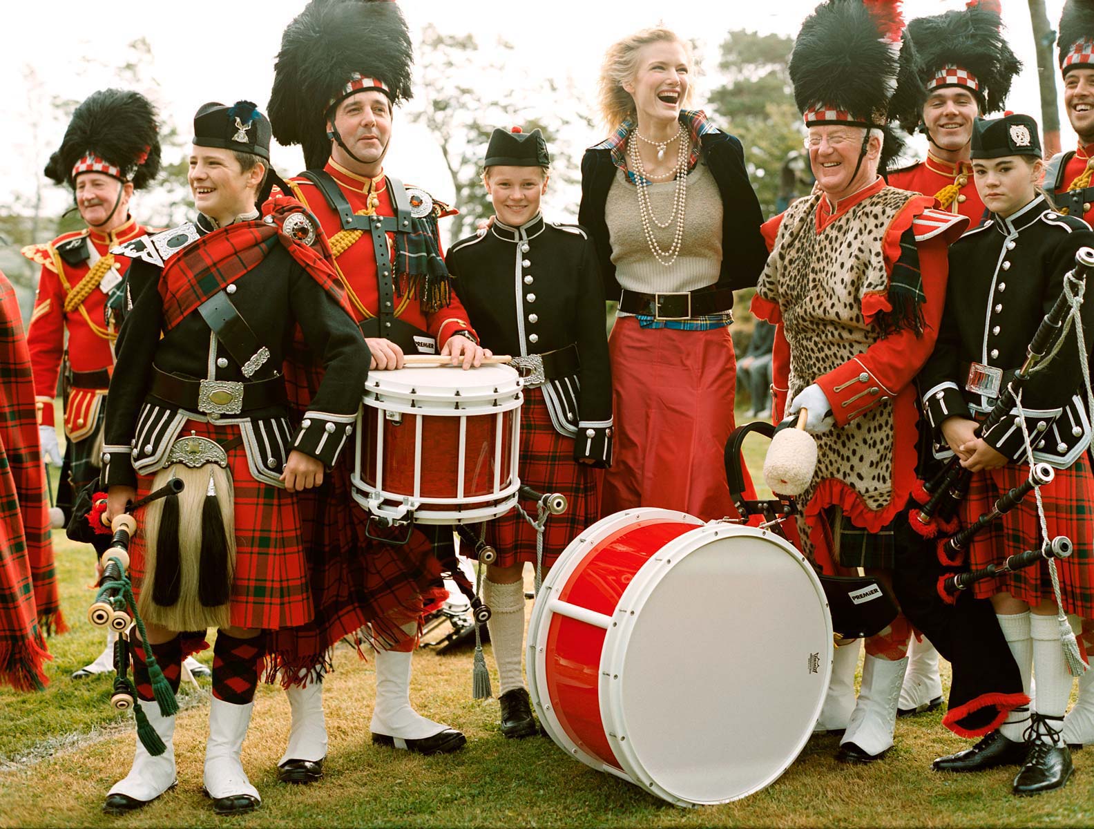 Outdoor gathering of Scottish pipe band members in traditional kilts, jackets, and hats, with drums, and a woman in modern clothing.