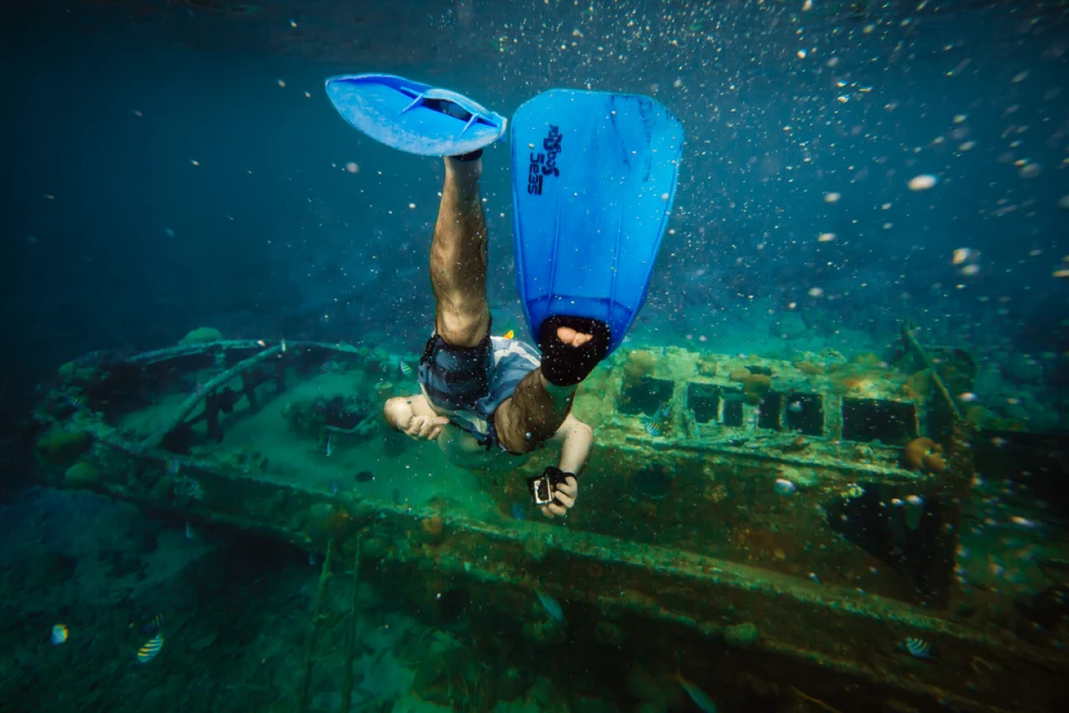 Underwater view of a scuba diver in blue flippers swimming near a sunken structure in the ocean, surrounded by marine life.