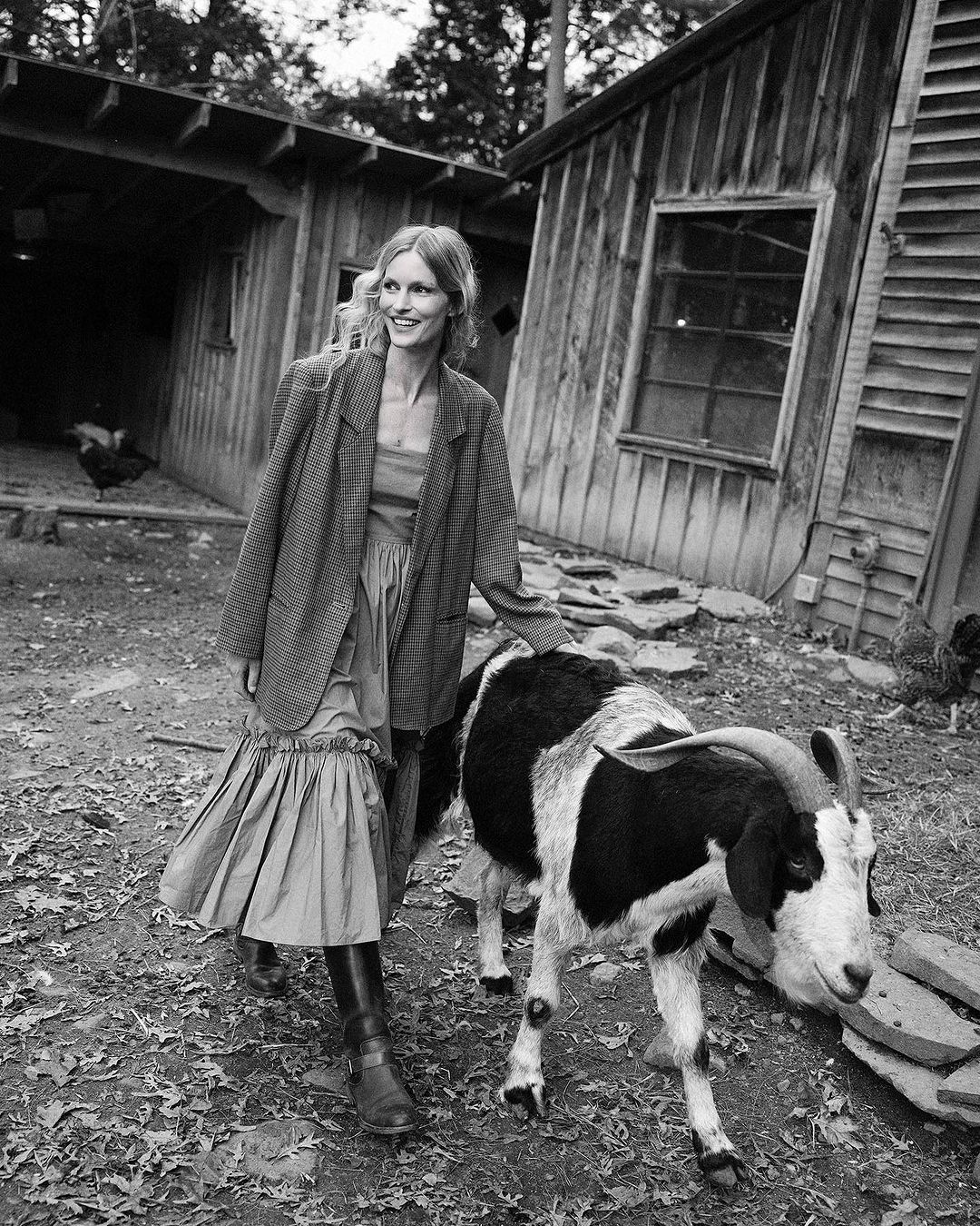 Monochrome photo of a smiling woman in a blazer and long skirt walking outdoors with a black and white goat near rustic wooden buildings.