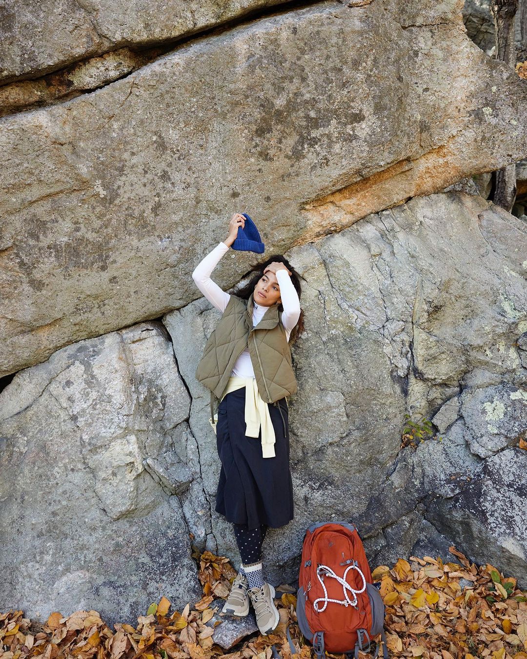 Woman in outdoor fashion, stands near rock face with backpack and fallen leaves. Wearing olive vest, skirt, white shirt, and beanie.