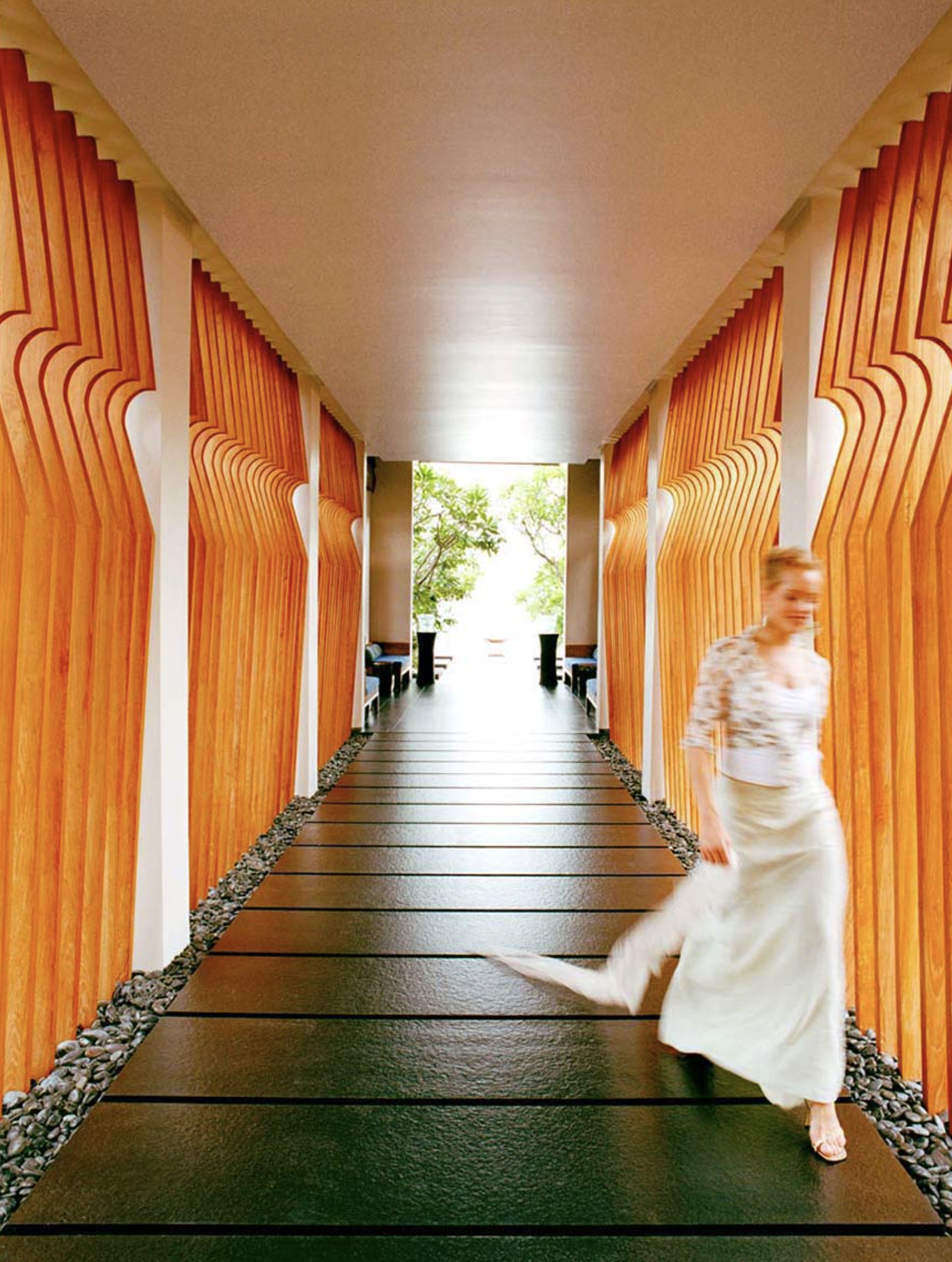 Woman in a flowing dress walks down a hallway lined with wooden panels and a black tiled floor towards a bright light.