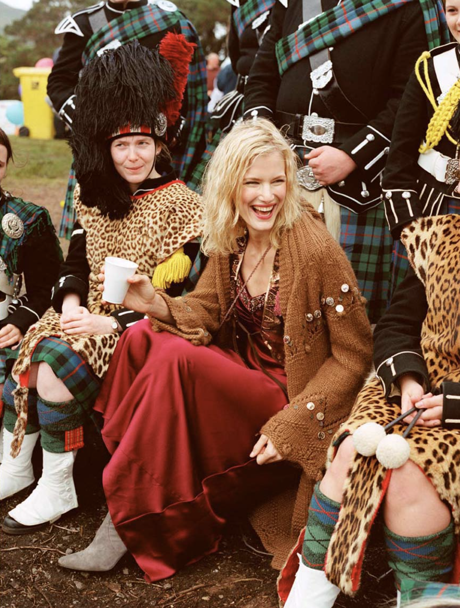 Smiling woman in red dress and brown cardigan sits among bagpipers in kilts and animal print during outdoor event, captured in medium shot.